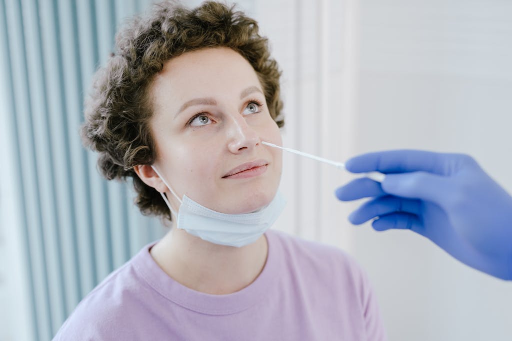 A woman in a medical facility receiving a nasal swab COVID-19 test, wearing a mask, smiling.